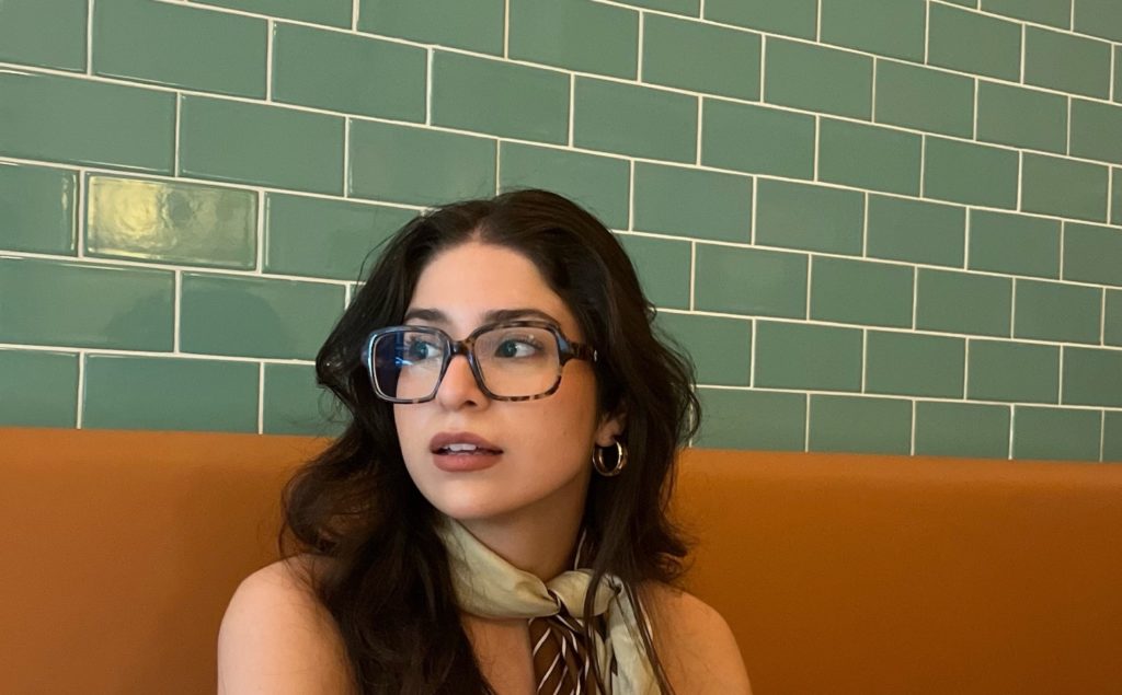 woman sitting at a booth wearing oversized glasses and a scarf tied around her neck