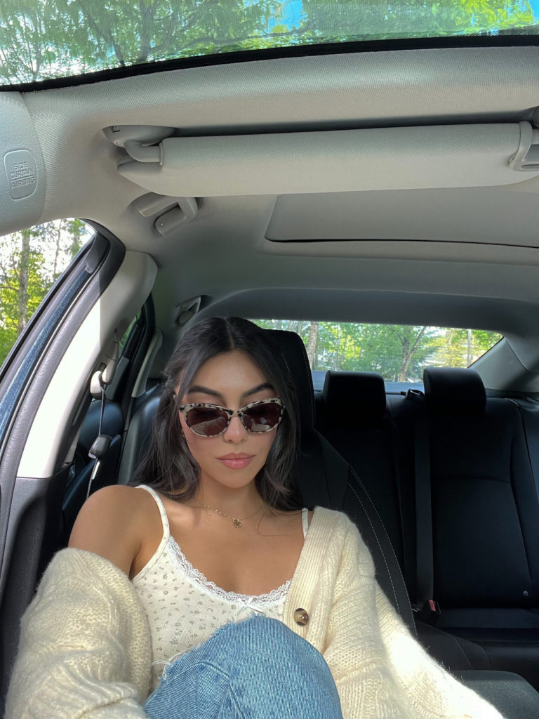 woman in a car wearing a sweater, floral top, jeans, and tortoiseshell sunglasses