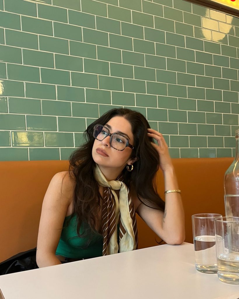 women sitting in a booth wearing oversized glasses and a scarf tied around her neck