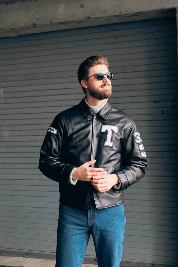 man wearing a leather jacket with a "T" on it and sunglasses, looking into the distance