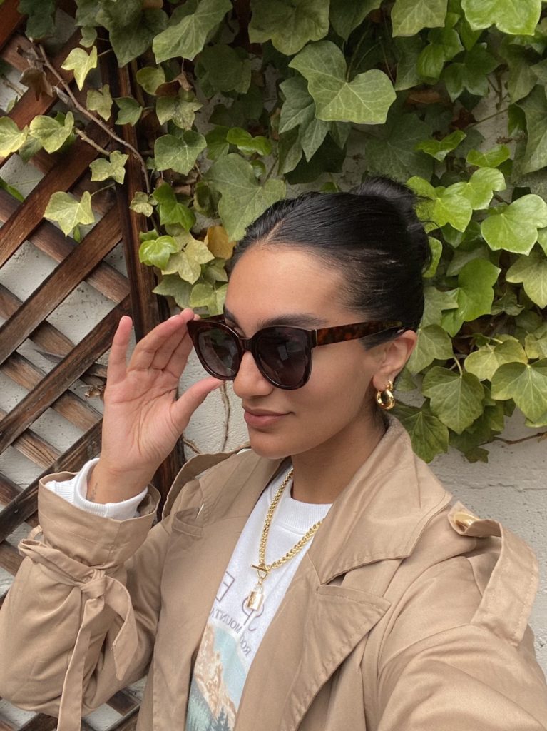 woman wearing oversized sunglasses and a beige trenchcoat in front of an ivy-covered wall
