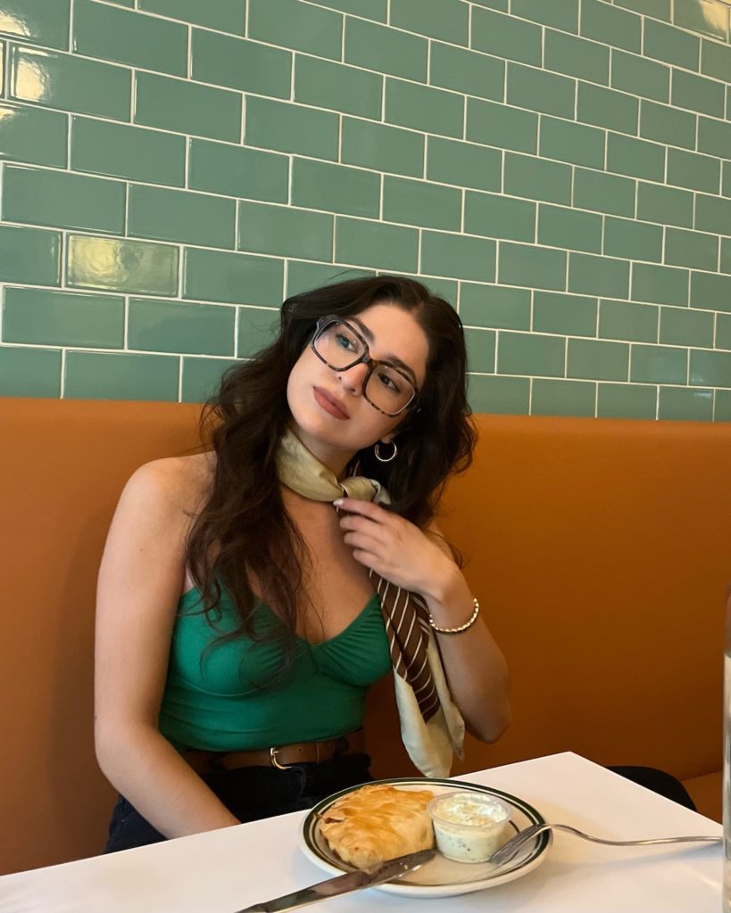 woman sitting in a diner booth with a green background wearing a scarf, green tank top and oversized glasses