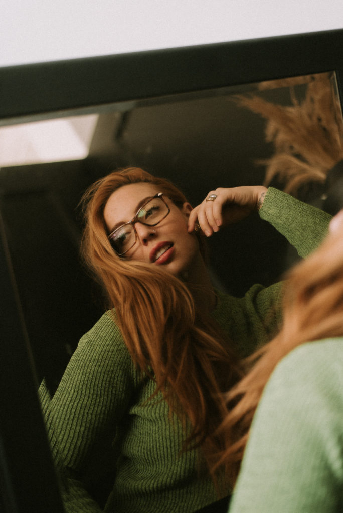 woman looking in the mirror wearing tortoiseshell glasses and a green sweater