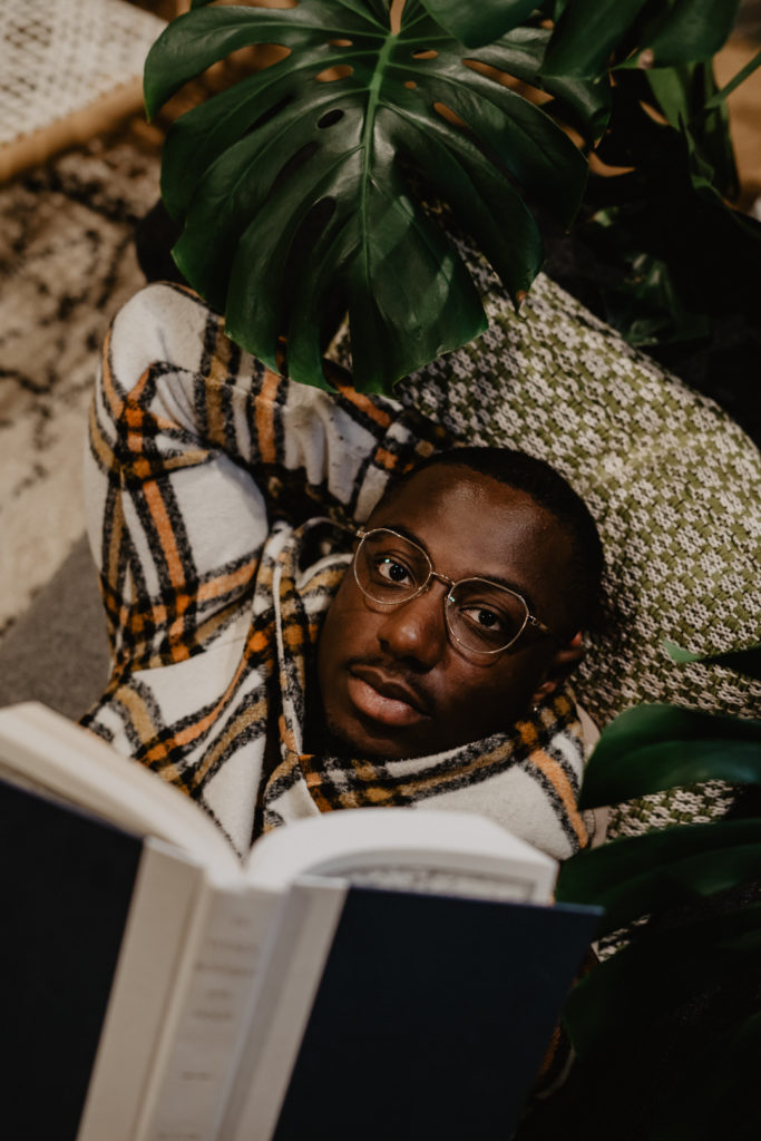 man lying on a couch reading a book wearing gold tortoiseshell glasses with leaves surrounding him