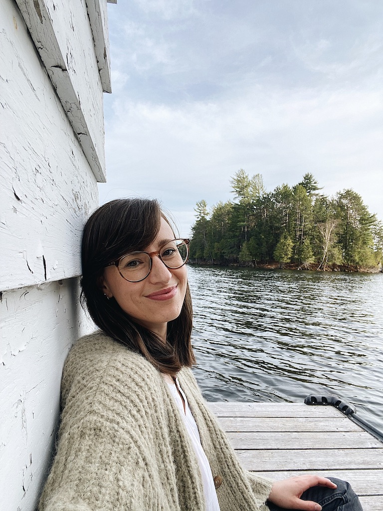 woman smiling sitting against a wall on a dock by a lake wearing round tortoiseshell glasses 