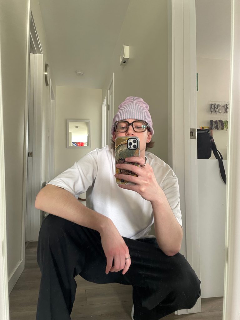 man crouching in front of a mirror taking a picture with his phone wearing oversized square tortoiseshell glasses 