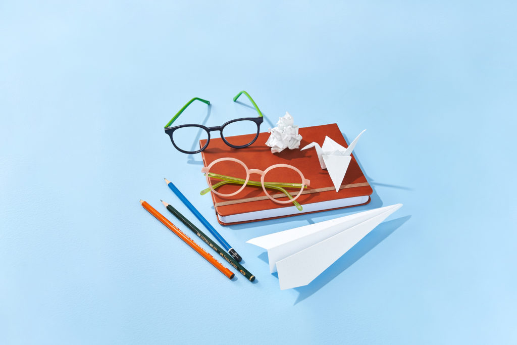 two pairs of colourful glasses sitting on top of a red notebook with a paper airplane and origami and pencils