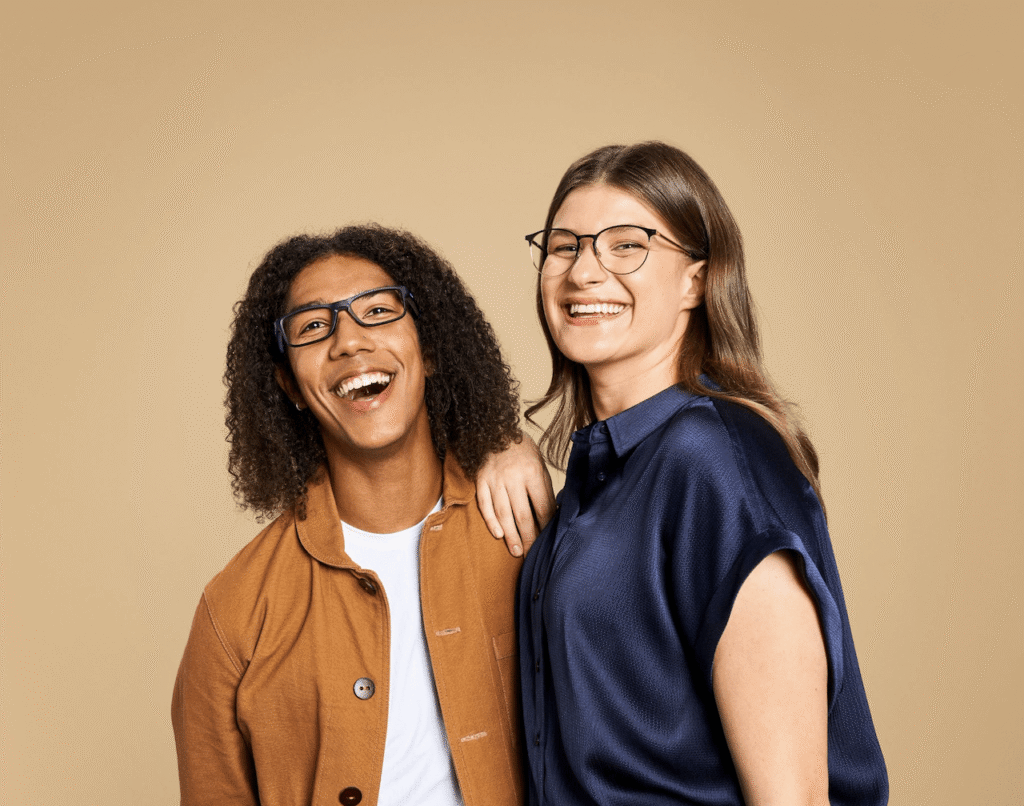 A smiling man and woman wearing eyeglasses in front of a tan background